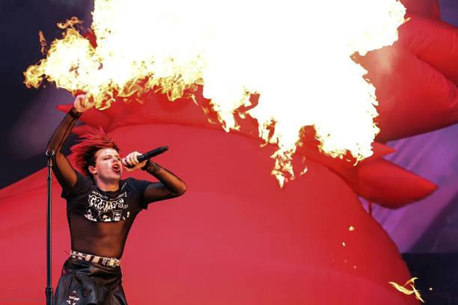A woman performing a fire breathing act with flames erupting from her mouth during a concert or performance on a stage with red and purple curtains.