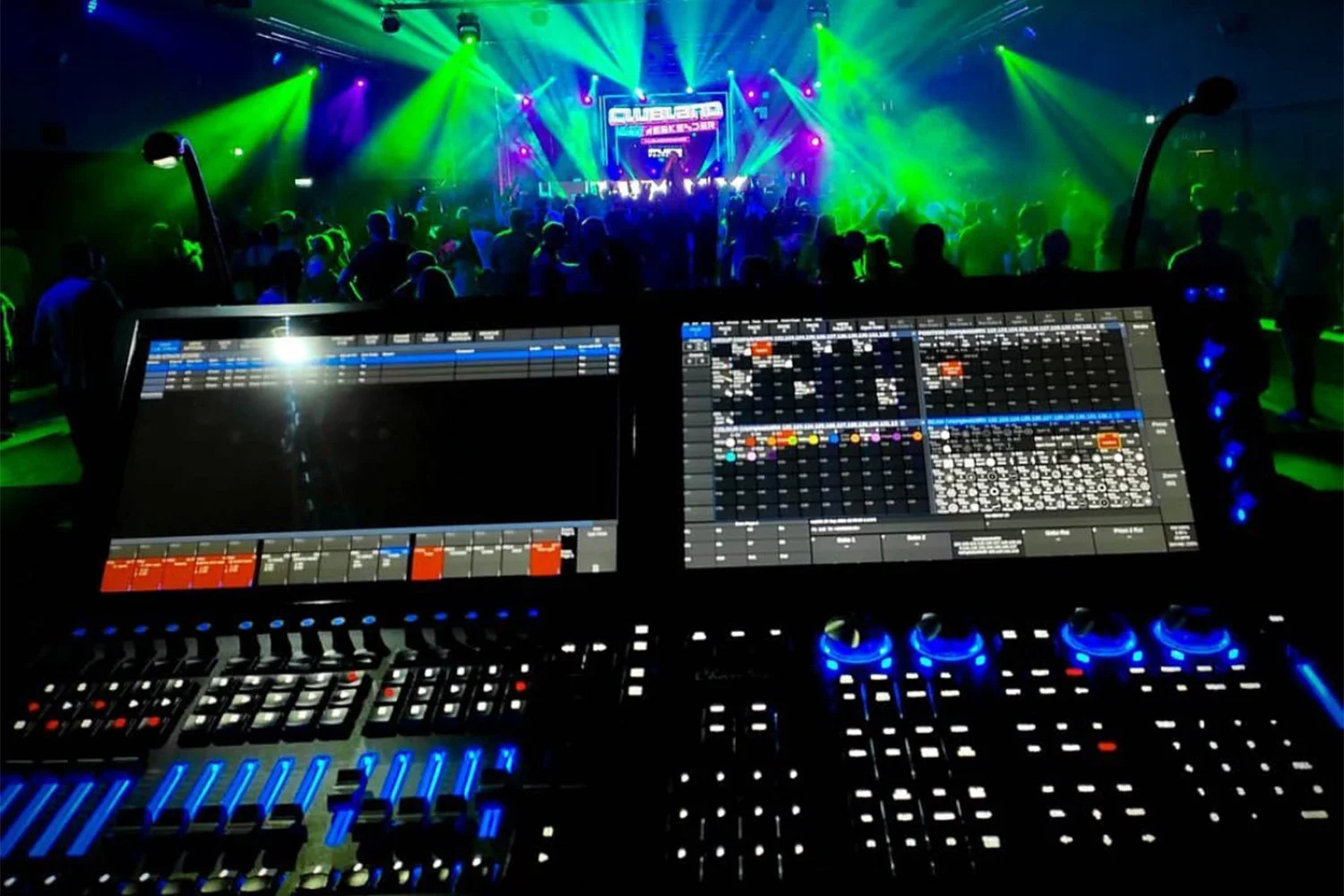 A DJ setup with two large screens displaying music controls, overlooking a dance floor with colorful lights and people dancing at a concert or nightclub.
