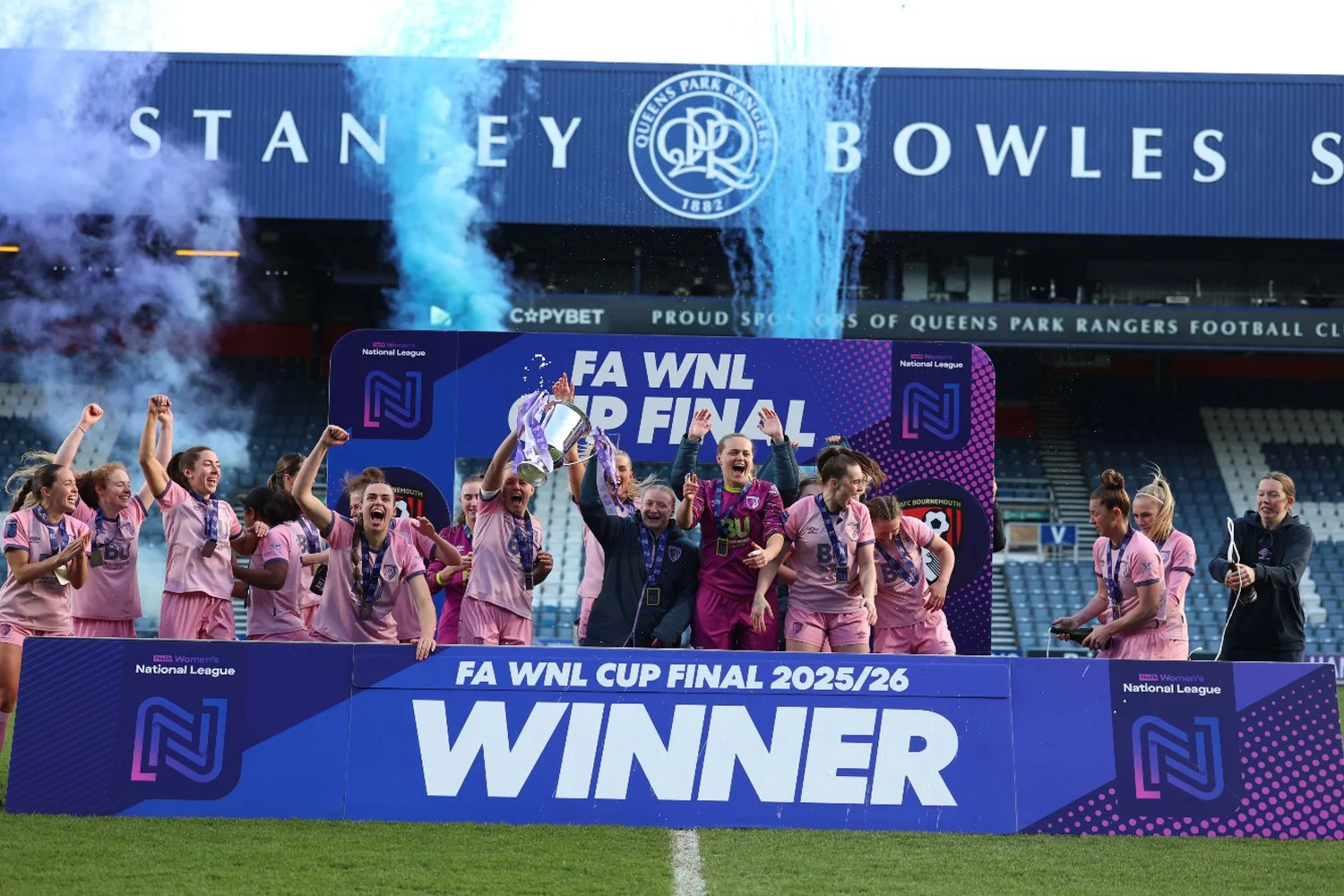 Women's soccer team celebrating winning the FA WNL Cup Final 2025/26 on the field at Queens Park Rangers football stadium. The players are in pink jerseys, some holding medals, with a large blue banner that says "WINNER" in front, and fireworks or smoke effects in the background.