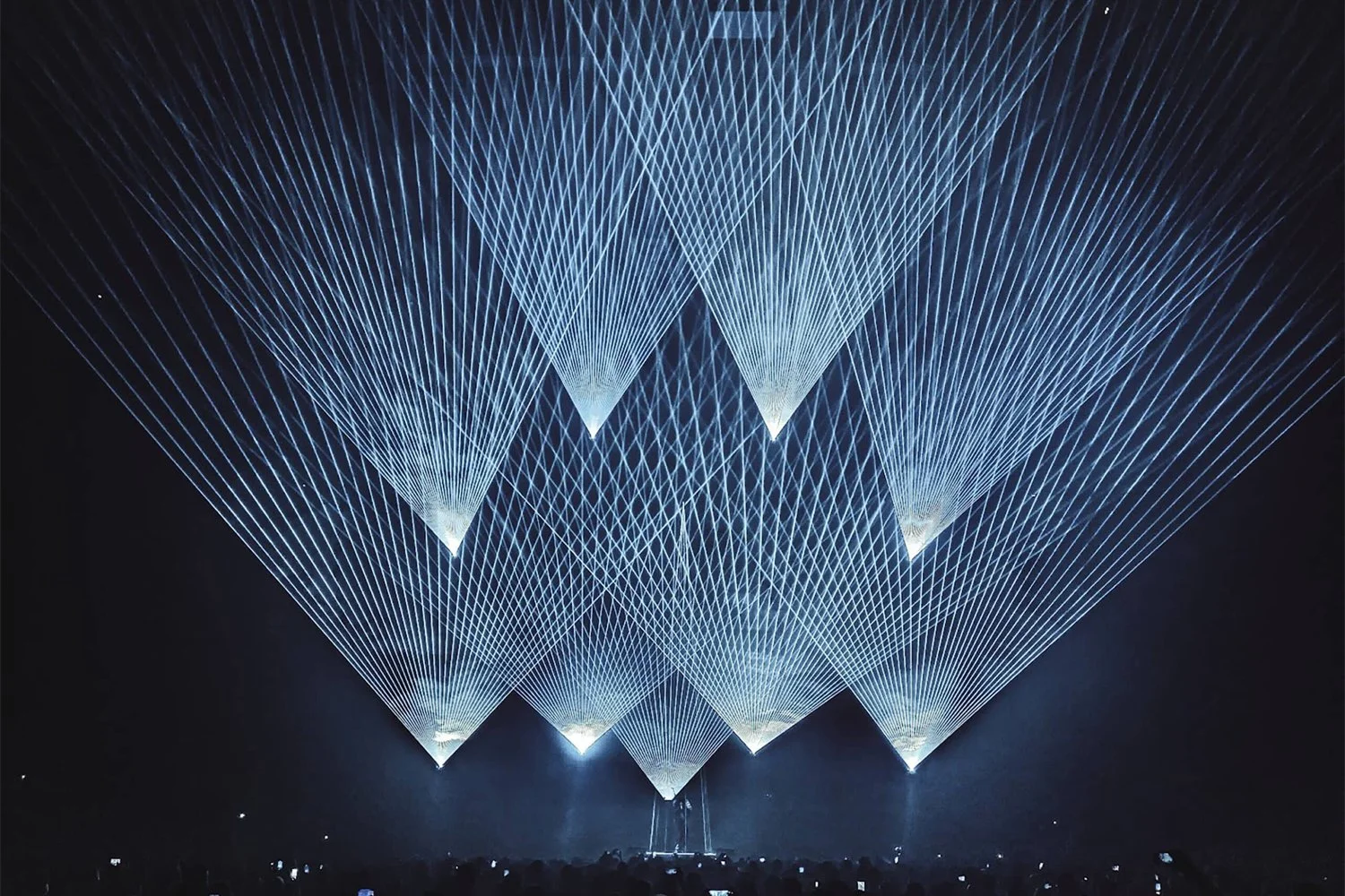 Laser light show with multiple blue and white beams forming geometric patterns above a crowd at a concert or event.