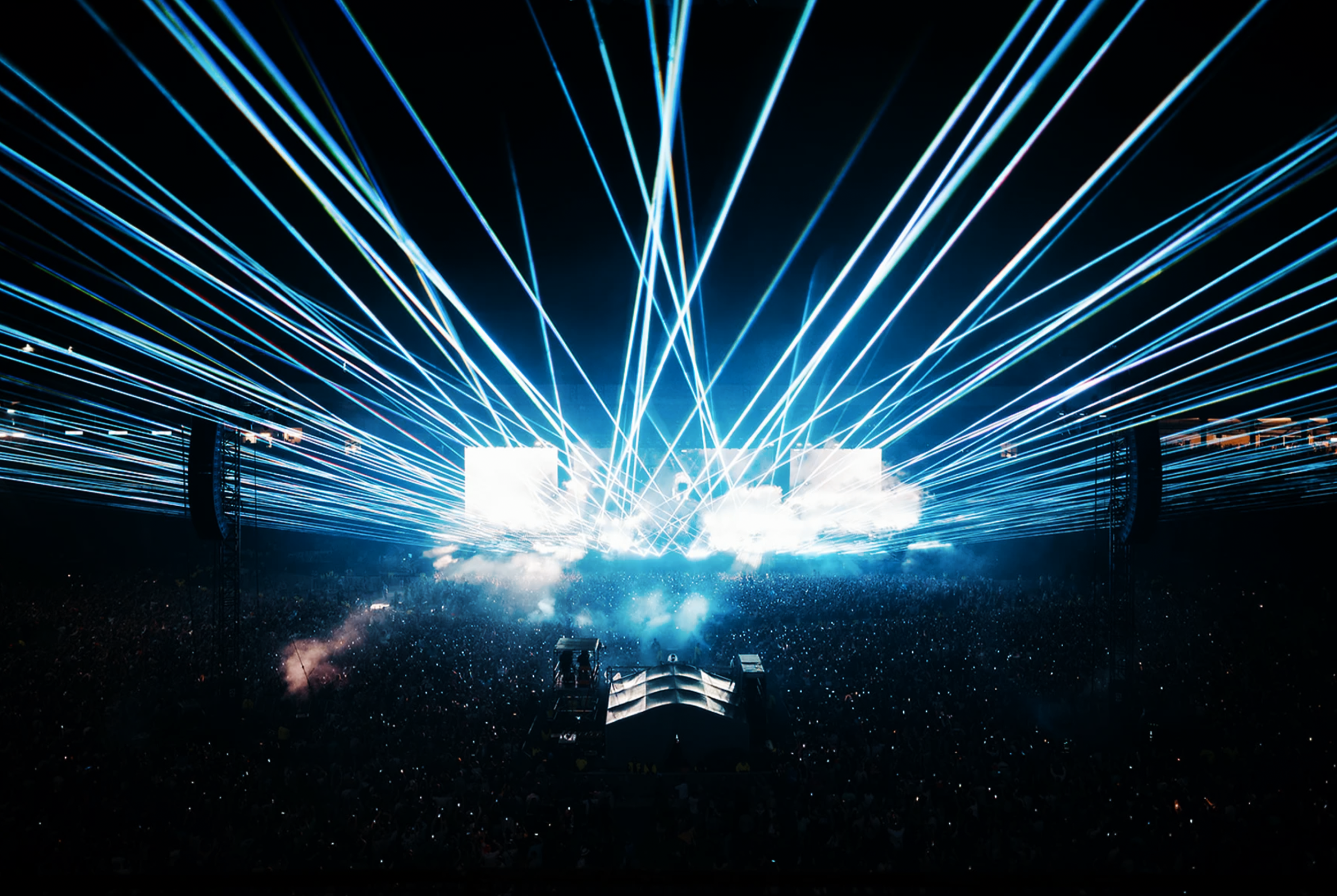 A concert or music festival stage with bright blue and white laser lights shooting across the dark sky, and an audience visible below.
