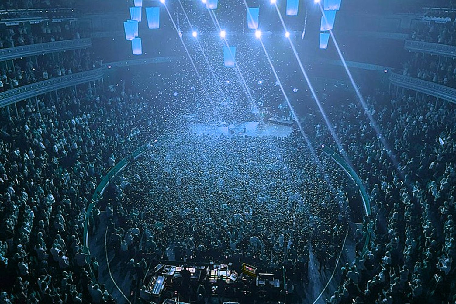 A large concert or event venue filled with a crowd of people. The stage is illuminated with bright lights and there are beams of light and confetti falling from above, creating a lively atmosphere.