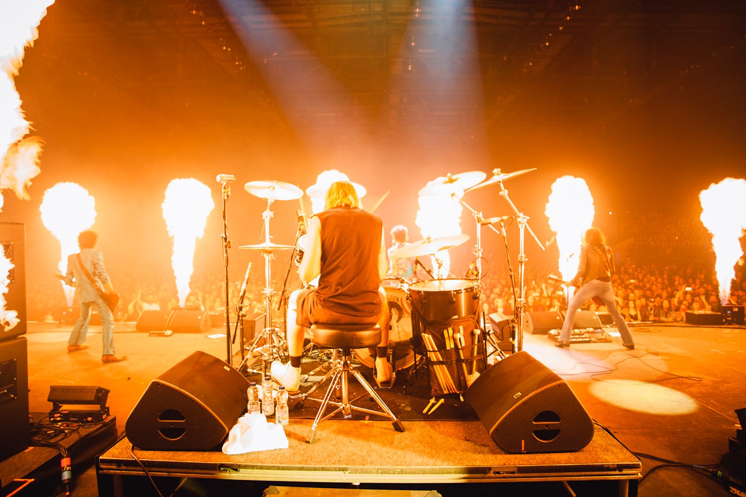 A live rock band performing on stage with pyrotechnics, including flames shooting upwards, and a drummer in the foreground.