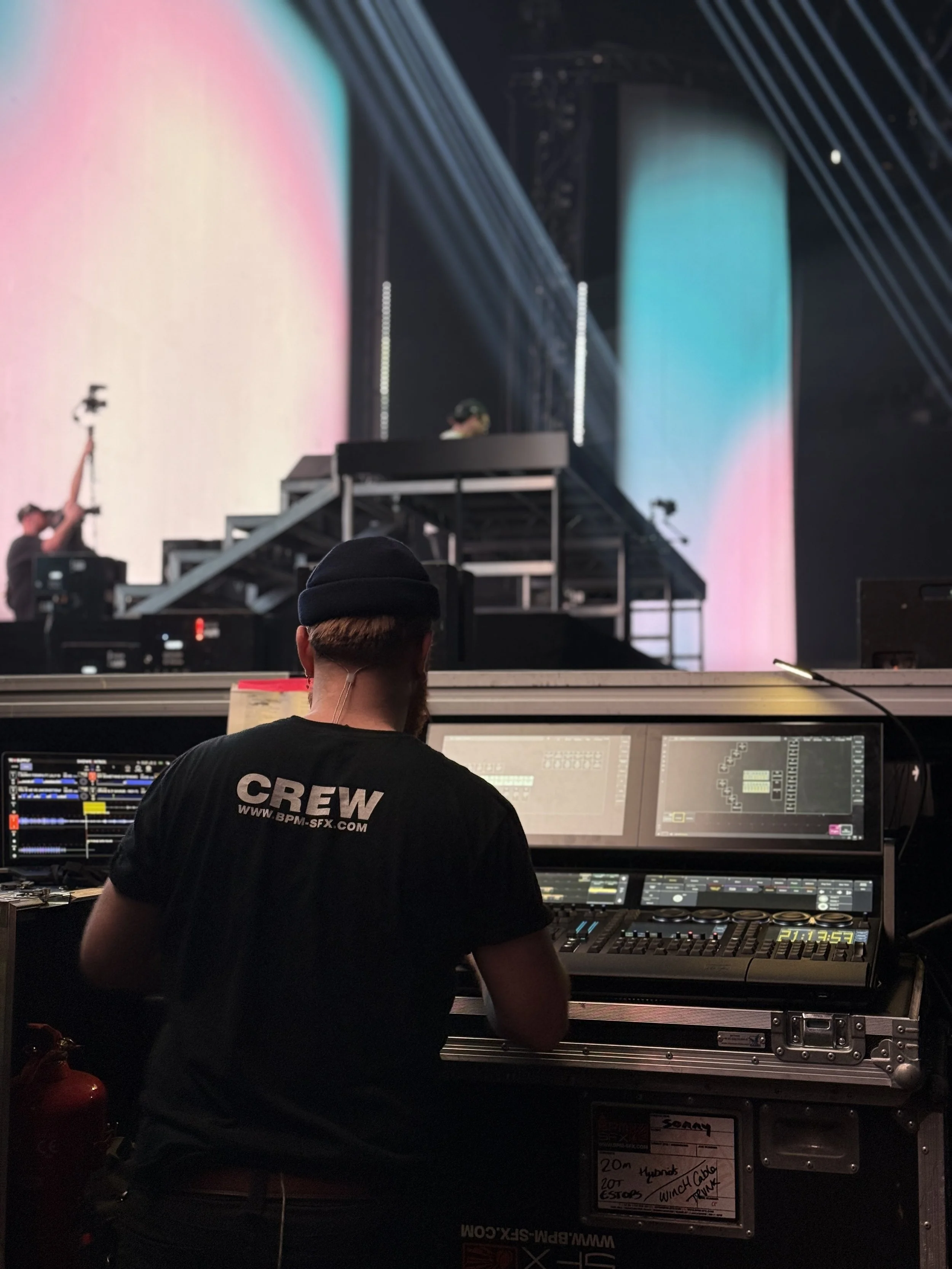 A person wearing a black cap and black t-shirt with 'CREW' written on the back, operating a professional lighting console at a concert or event. The background shows a stage with large LED screens and lighting effects.