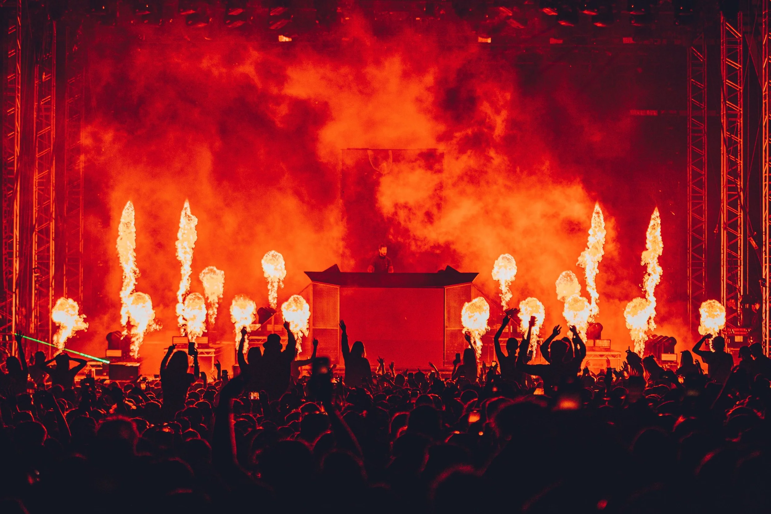 People dancing and enjoying a concert with bright fireworks and flames on stage, with the DJ and stage lights creating an energetic atmosphere.