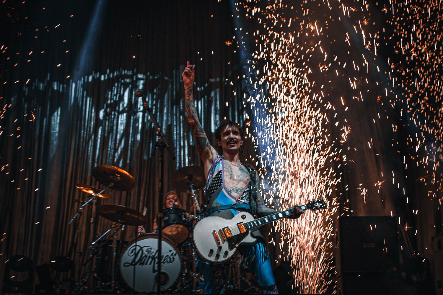 A male guitarist with tattoos performs on stage with a drummer behind him. Sparks are flying in the background as part of a concert show.