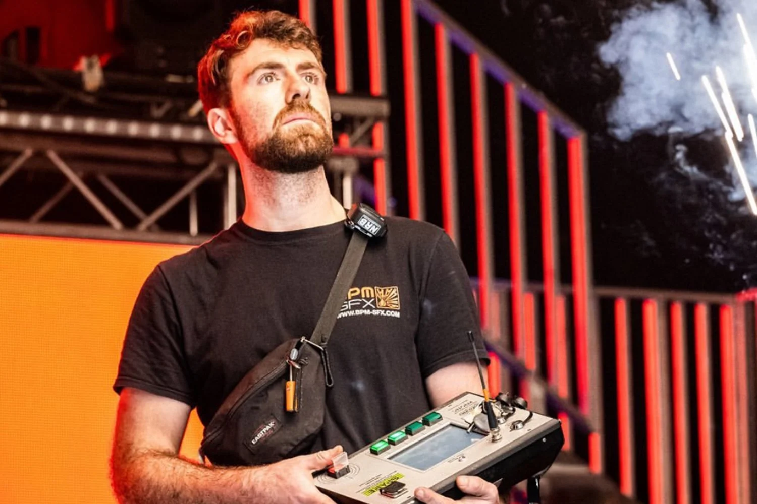 Man with a beard and short brown hair holding a video camera control in a dark stage setting with red vertical light strips in the background.