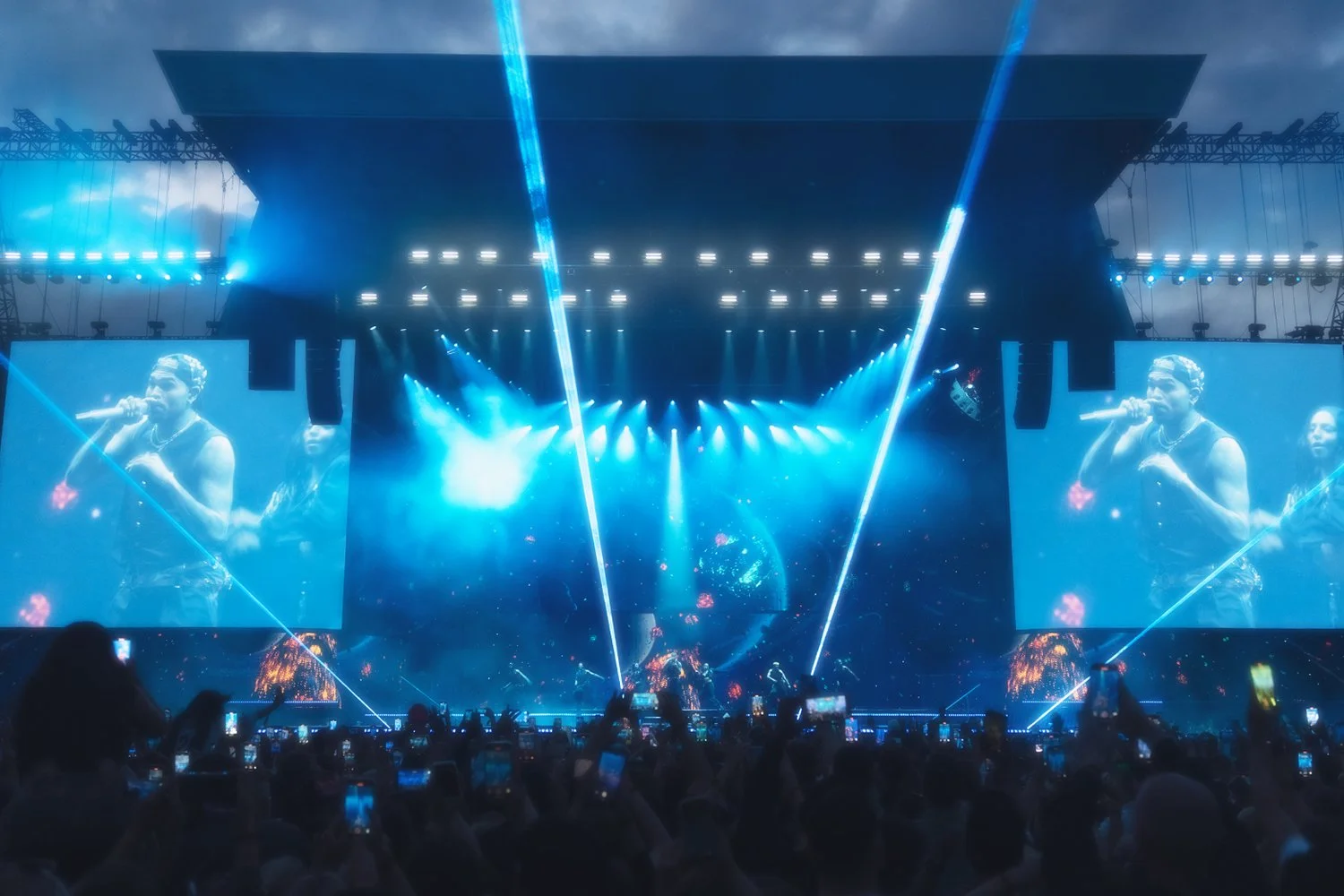 A concert stage illuminated with blue lighting, featuring large screens showing performers, with laser lights and an audience capturing the show with smartphones.