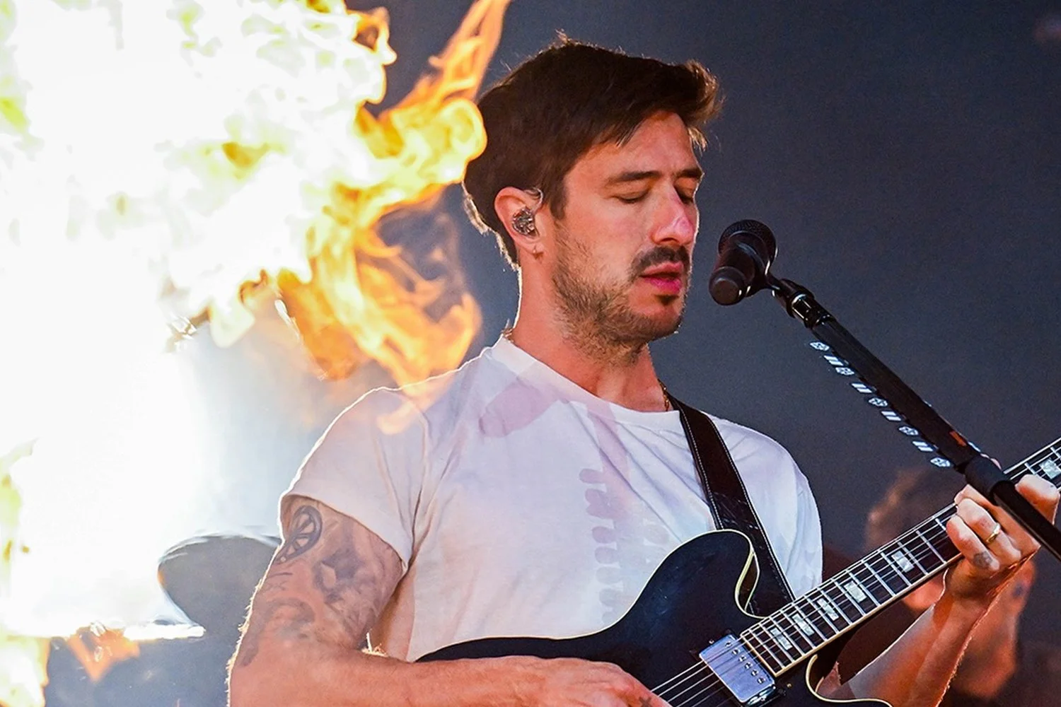 A man with dark hair and tattoos on his arms playing an electric guitar with a microphone in front of him during a performance, with flames and bright lights in the background.