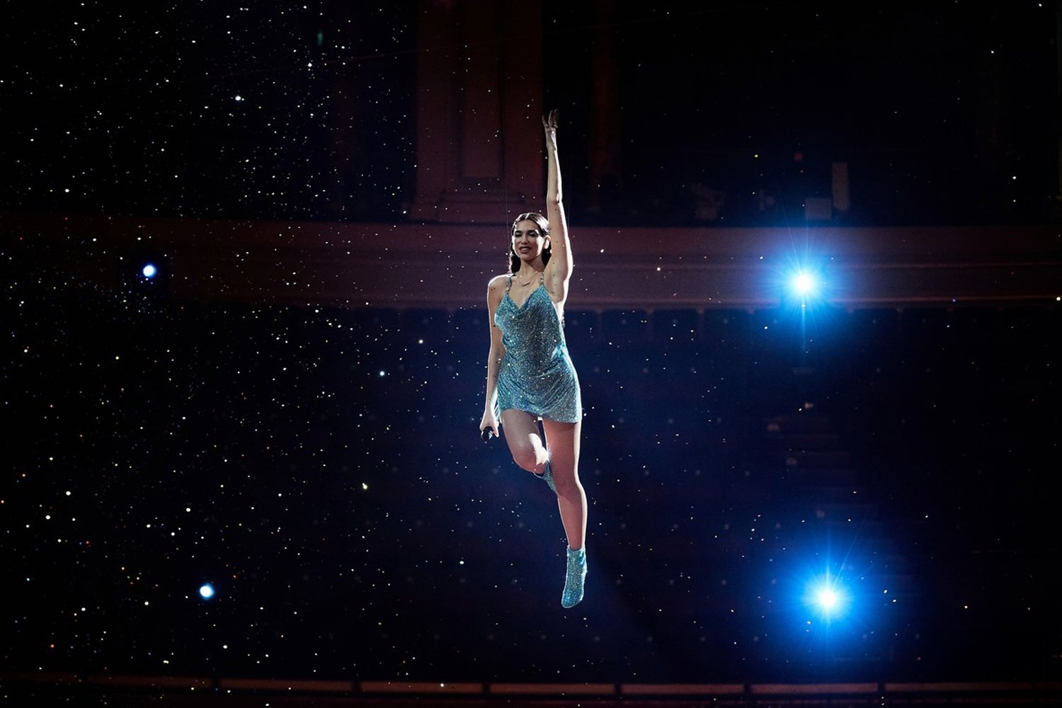 A female performer in a sparkly blue dress and matching boots arches backwards with one arm raised, suspended in mid-air against a dark backdrop with star-like lights and bright blue stage lights.