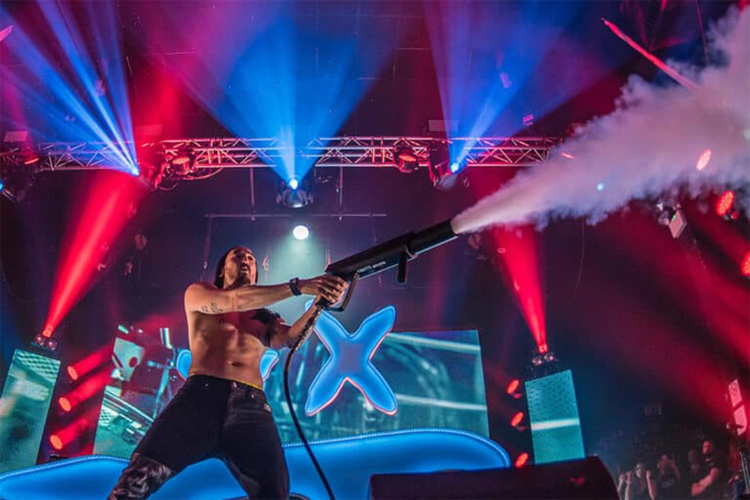 A shirtless man on a stage aims a fog blasting device at the audience with colorful lights and a large star-shaped screen in the background.