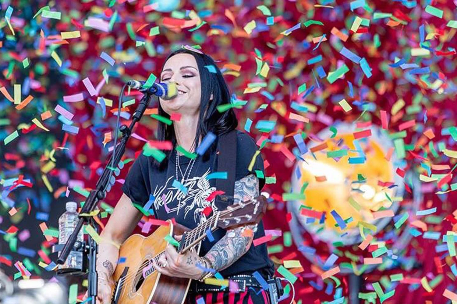Woman with black hair and tattoos playing an acoustic guitar and singing into a microphone surrounded by colorful confetti, with a blurred background featuring a large candle or similar object.