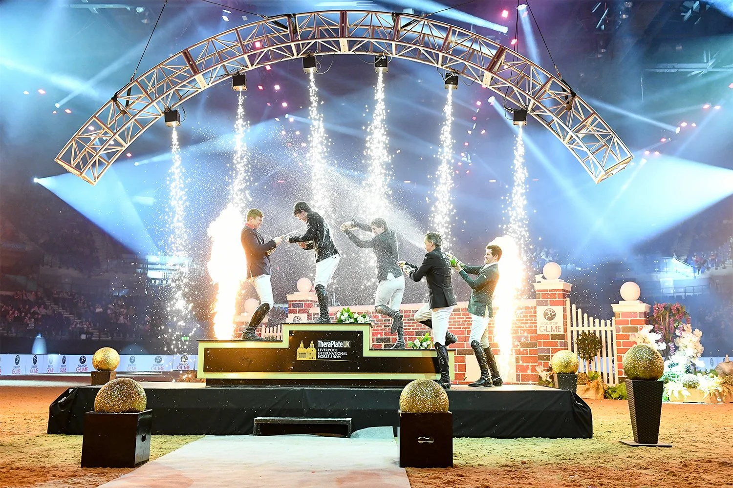Celebration of event with five men dressed in equestrian attire on a podium, spraying fireworks, and water fountains at an indoor arena with decorated setting and an audience in the background.