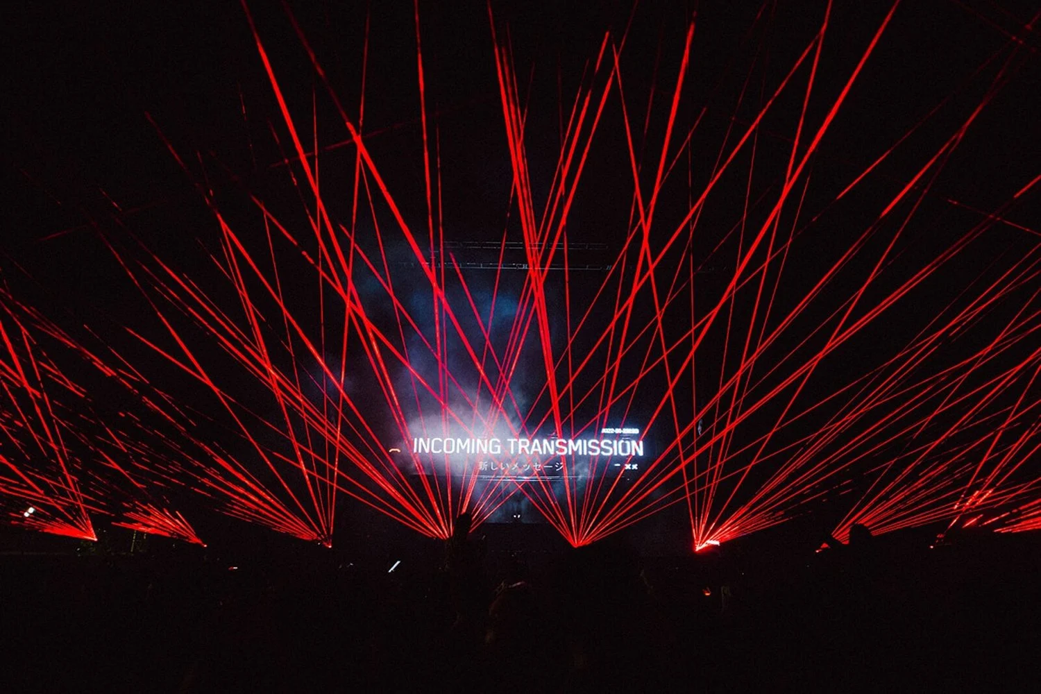 A dark concert or event venue illuminated by red laser lights projecting downward, with a central screen displaying the text 'INCOMING TRANSMISSION' and smoke or fog effects.