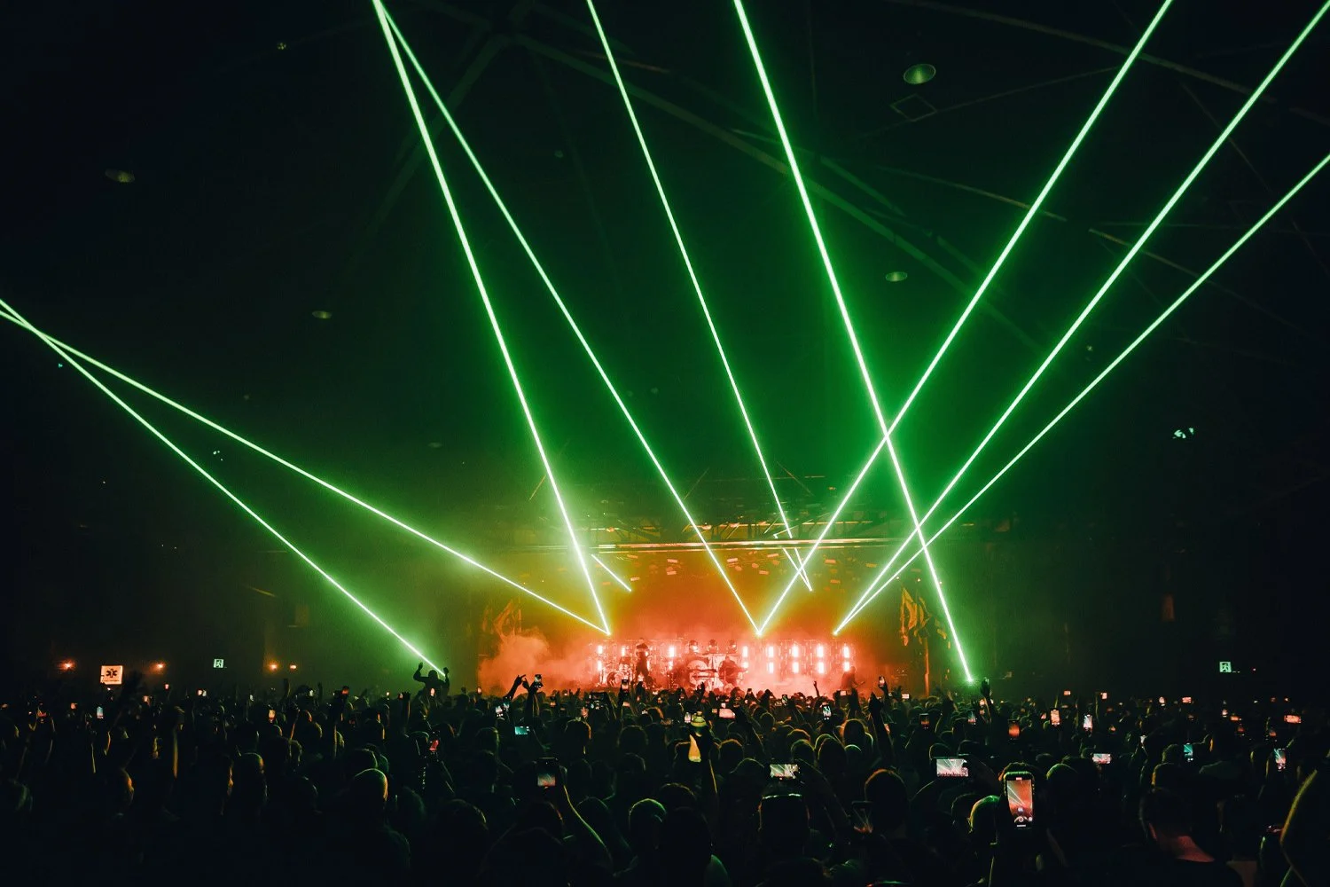 Concert scene with green laser lights and a large crowd of fans taking photos and videos.