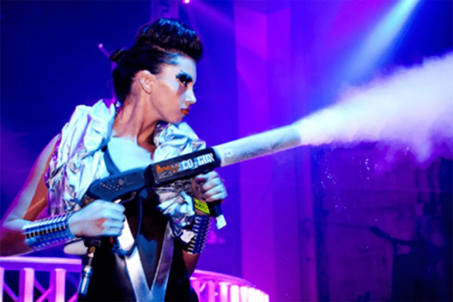 A person dressed in futuristic fashion with colorful makeup is using a paintball or foam gun that shoots foam or paint in an indoor setting with purple and blue lighting.