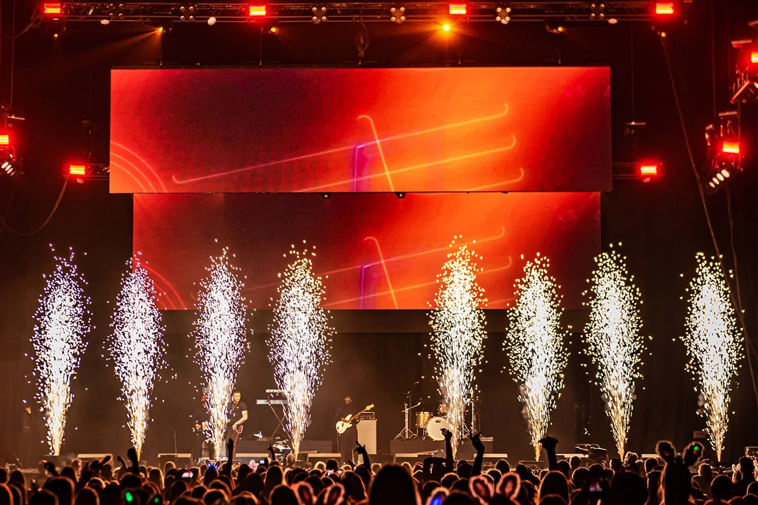 A concert stage with fireworks and pyrotechnics, band performing, large LED screens displaying abstract graphics, and a crowd of people watching.