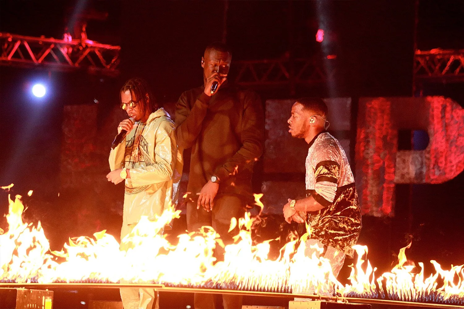 Three men are singing on a stage with a line of fire in front of them, lit by stage lights.