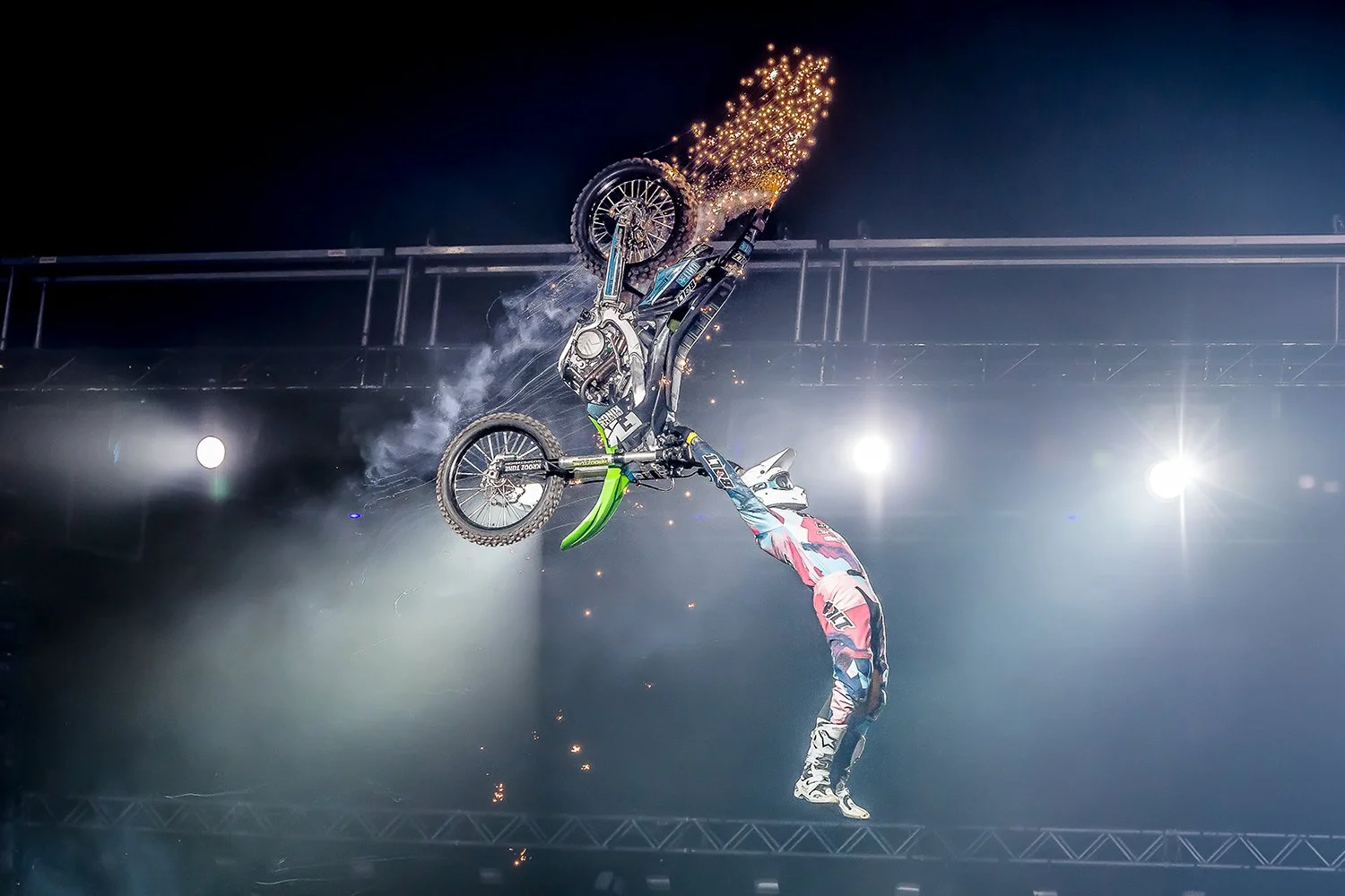 Motorcycle stunt performer mid-air during a stunt, with sparks and smoke, wearing colorful gear, performing a flip against a dark background with bright lights.
