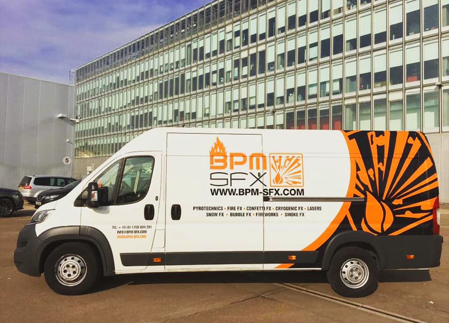 A white service van with orange and black graphics and text parked in front of a modern multi-story glass building. The van's side displays the logo and website for BPM SFX, a company specializing in pyrotechnics and special effects.