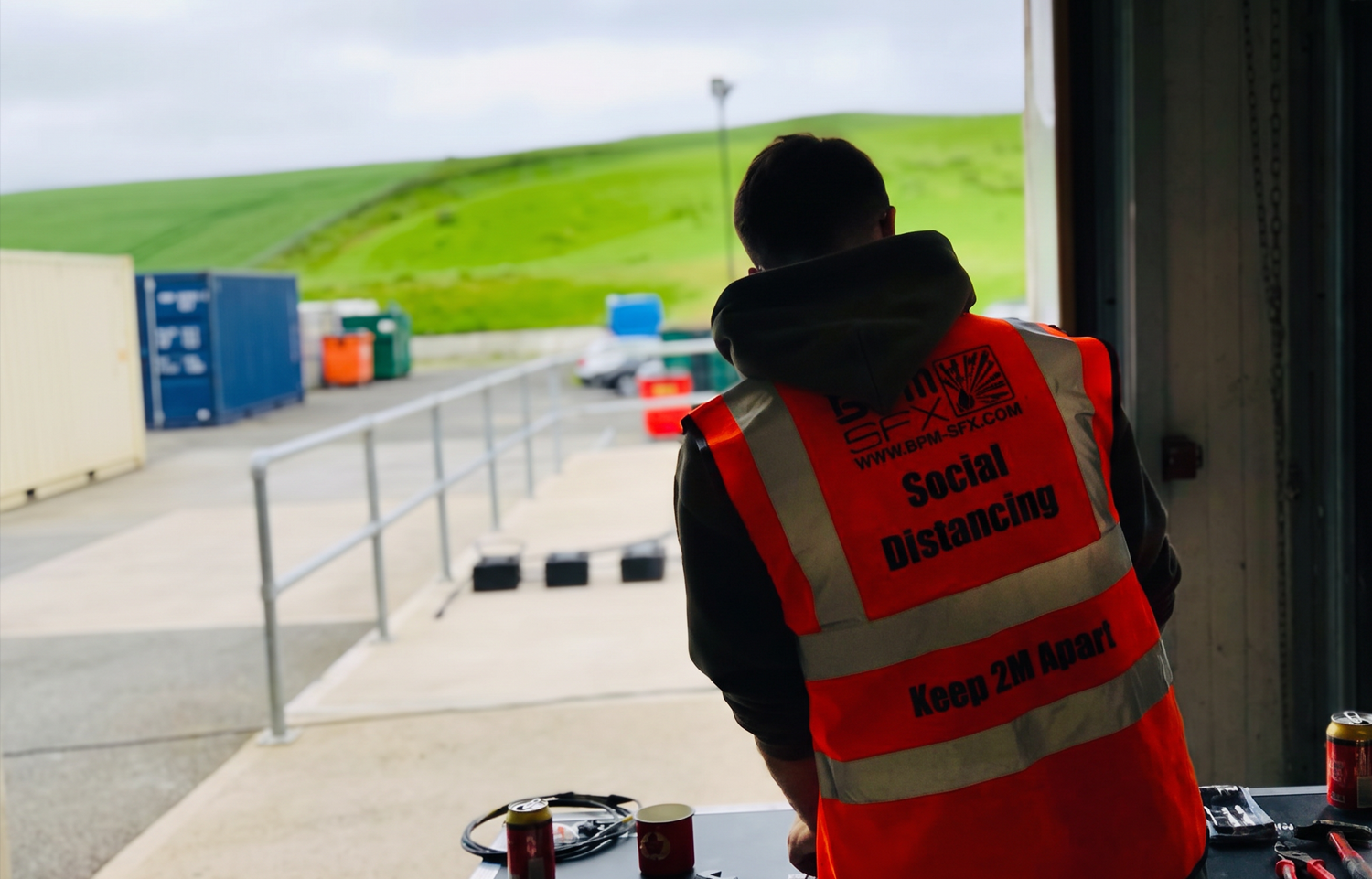 Person wearing a bright orange safety vest with social distancing instructions, standing indoors and looking out towards an outdoor parking area with containers, cars, and a green hill in the background.