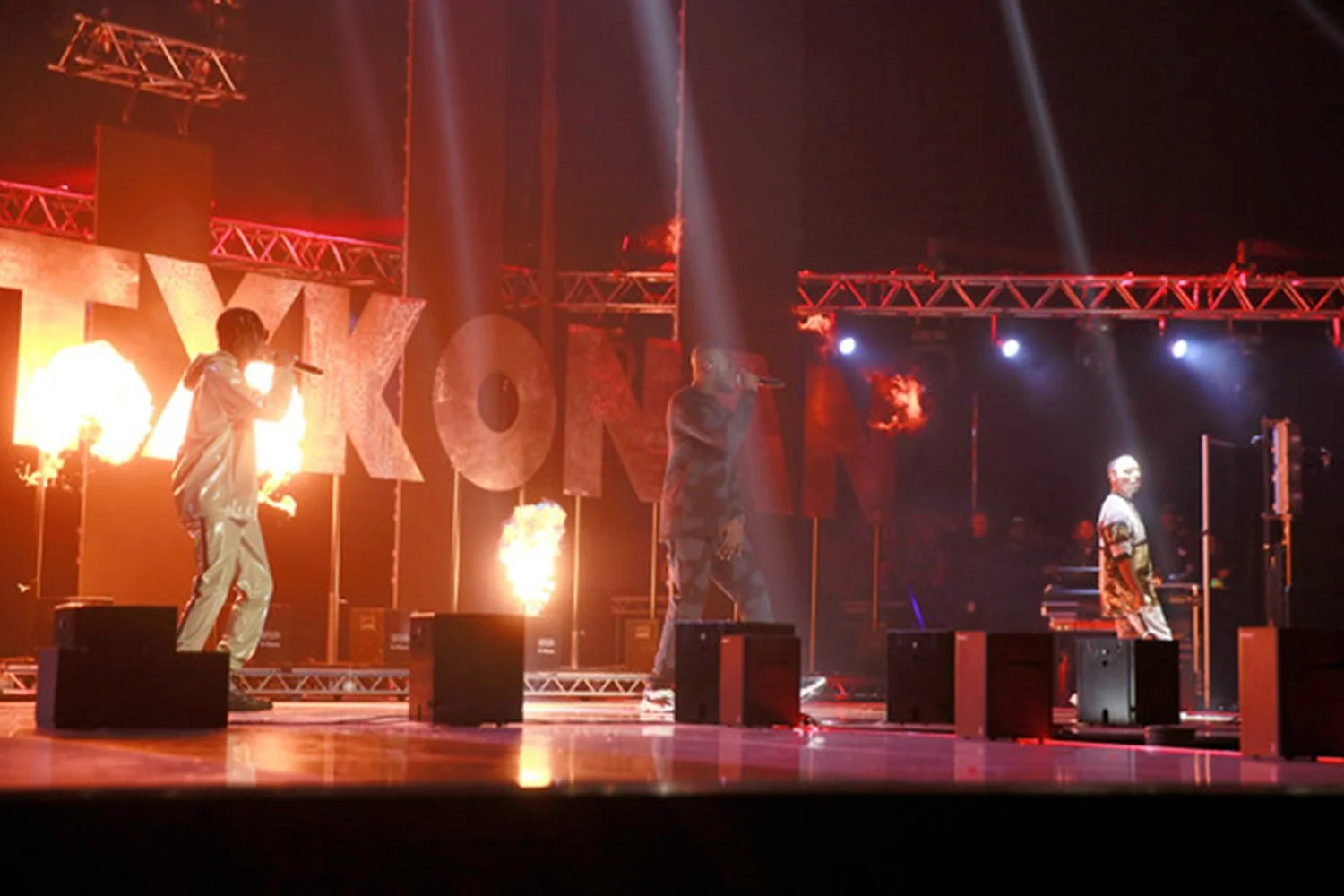 Three performers on stage with fire effects and large illuminated 'TOKYO' sign behind them, performing at a concert or event.