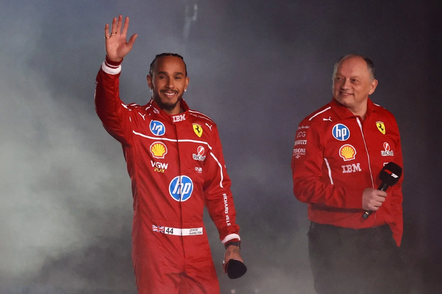 Two men wearing red Ferrari racing jackets, one waving and the other holding a microphone, standing on a stage.