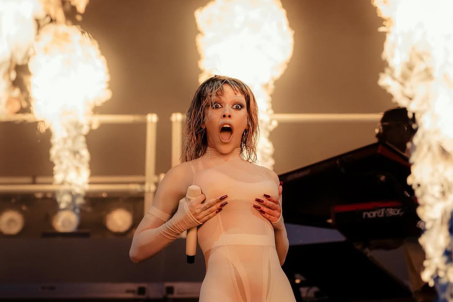 A woman with wet hair, wearing a nude-colored dress and gloves, with a surprised or shocked expression, standing with her hands on her chest in front of flames and a piano.
