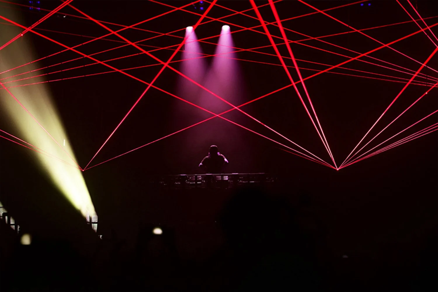 A DJ performing on stage with laser light effects, purple and yellow spotlights, and an audience in the foreground.