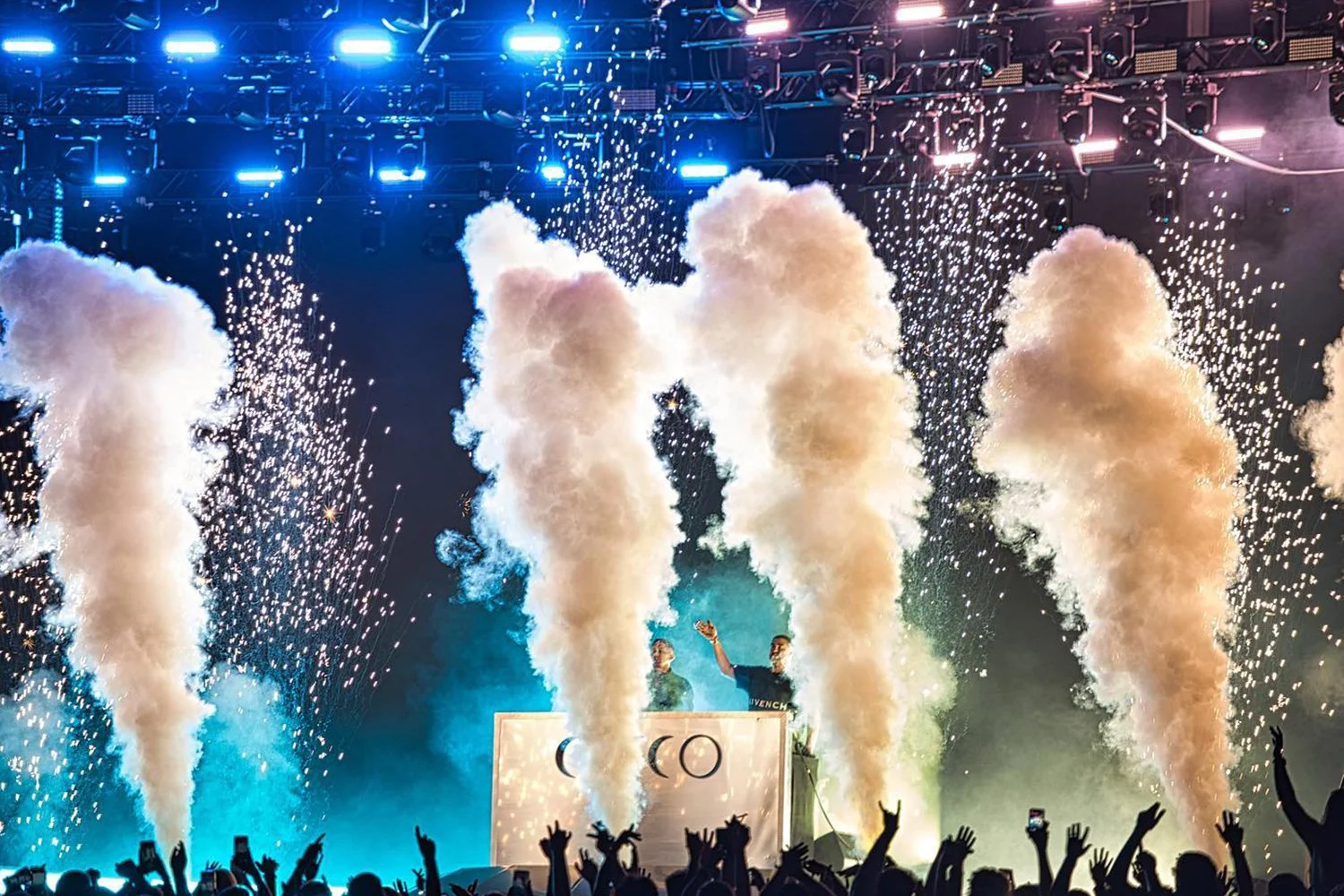 A live concert with pyrotechnics, smoke effects, blue stage lighting, and an audience raising their hands.