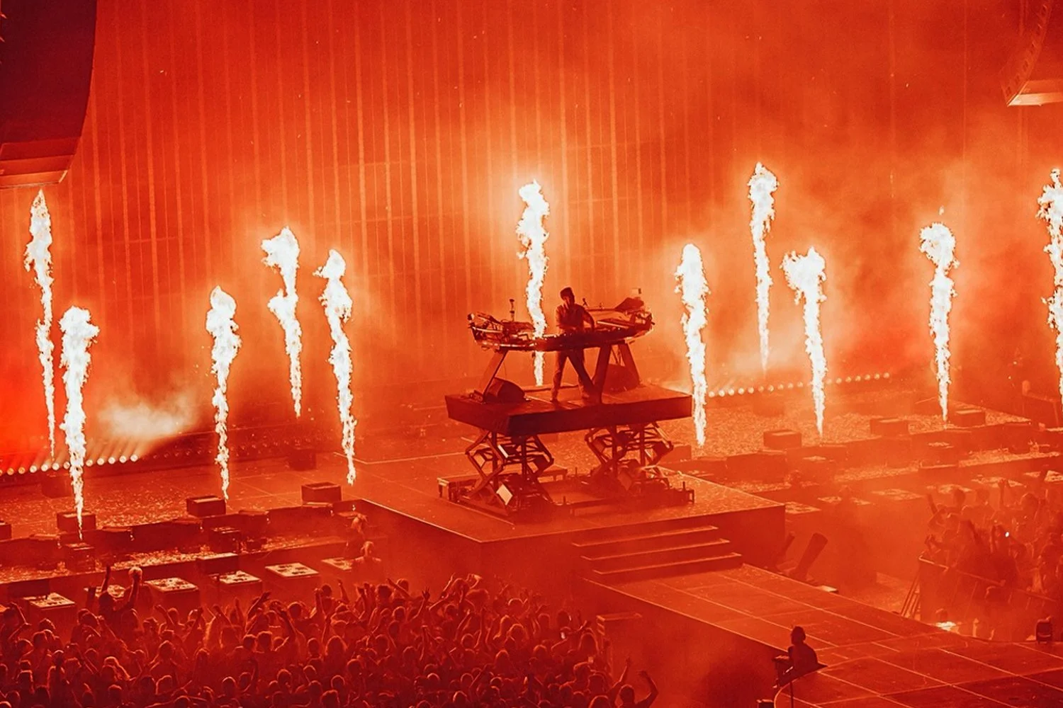 A DJ performs on an elevated stage with fire effects shooting upward behind them at a concert or nightclub, with a large crowd in front enjoying the show.