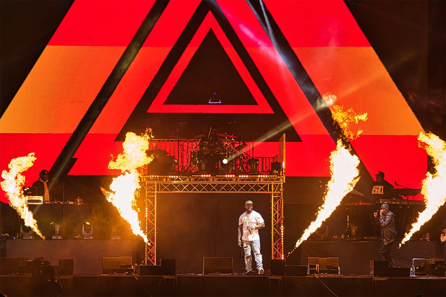 A live musical performance on stage with pyrotechnics. A singer stands at the front, with a band and DJ behind them. The stage features a large digital screen displaying red and black geometric triangles.
