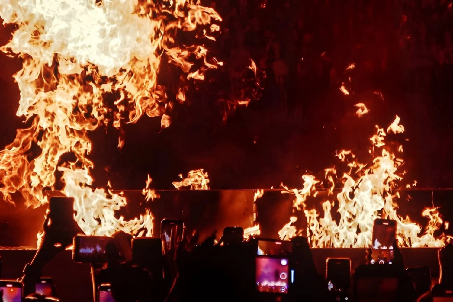 Large fire with bright flames at night, with several people in the foreground capturing the scene on their phones.