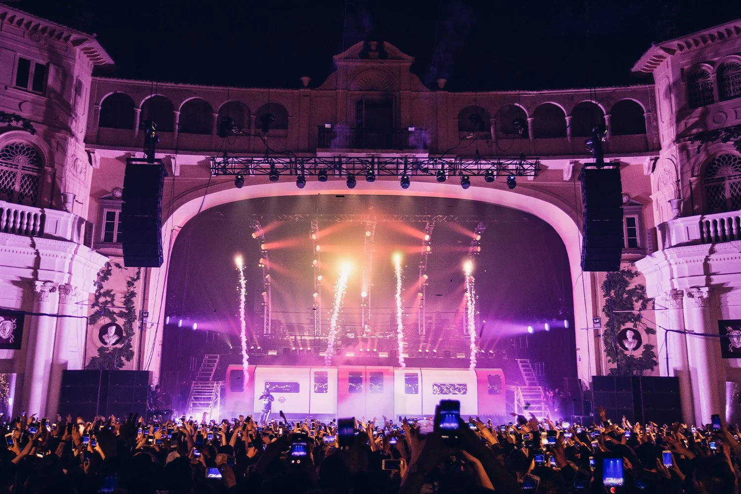 A concert stage with bright lights, pyrotechnics, and a crowd of people taking photos at night.
