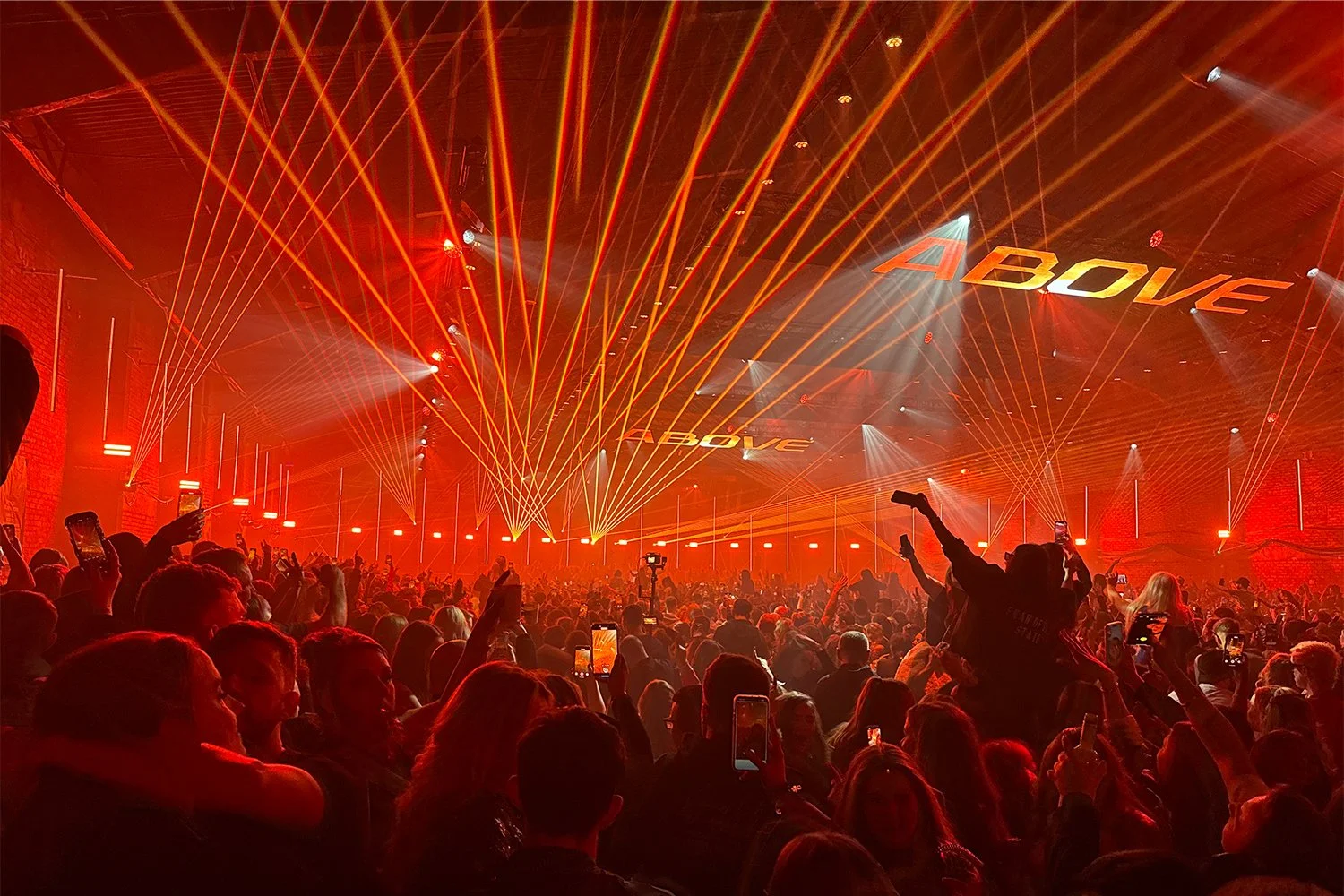 A large crowd at a concert or event with bright red lights and laser beams, some people are holding up their phones to take pictures, and the word 'ABOVE' is projected on the ceiling.
