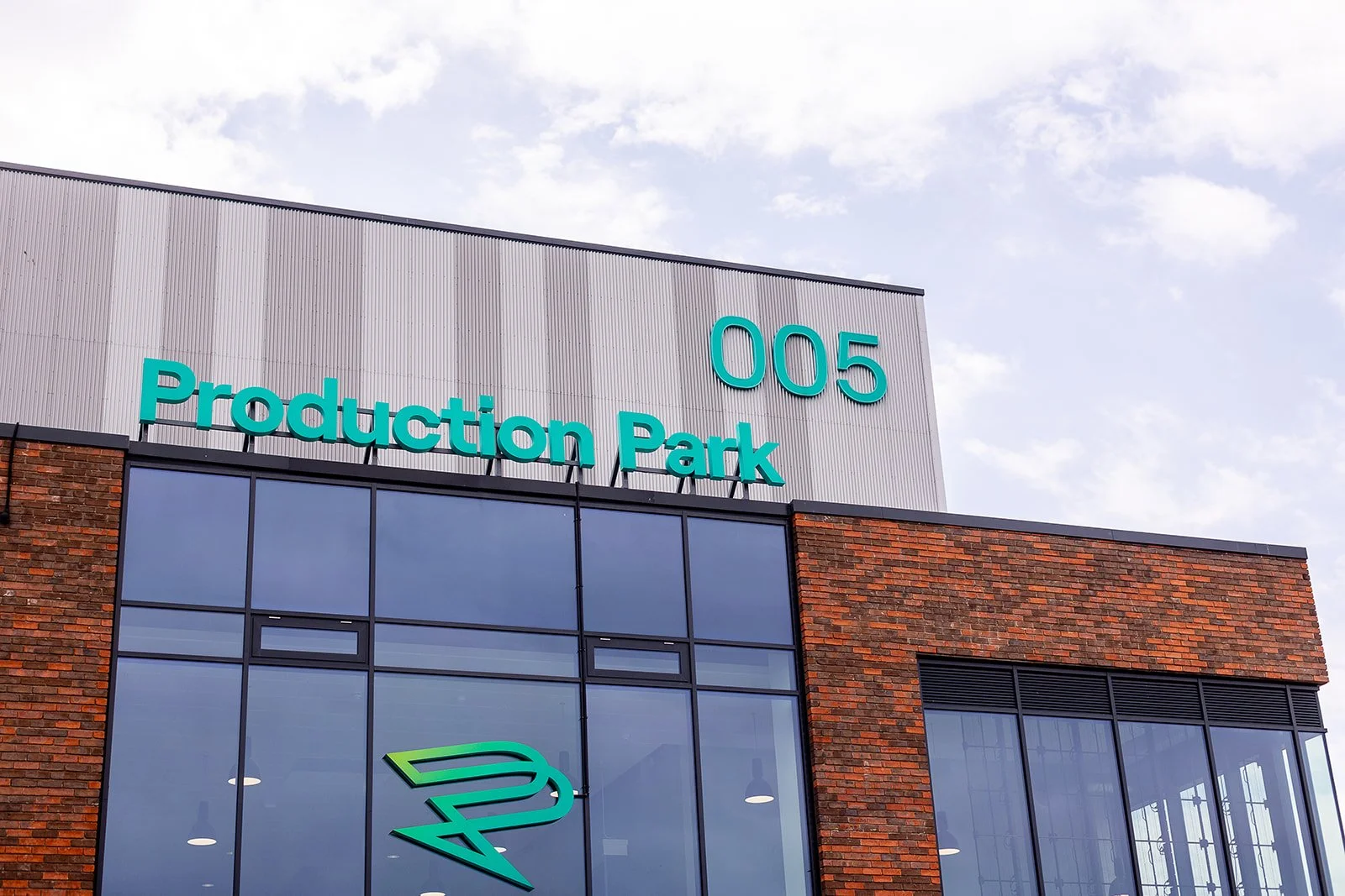 Exterior of a modern building with large glass windows and brick walls, displaying the address 005 and the sign 'Production Park' in teal letters, under a partly cloudy sky.
