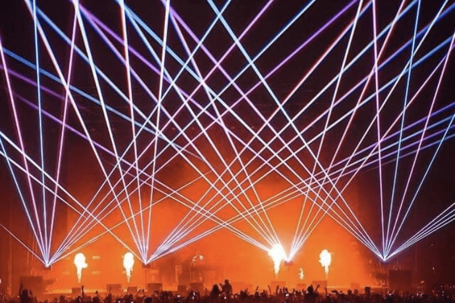 A concert stage with bright laser lights in blue, pink, and purple crossing overhead, with smoke and pyrotechnic effects, and silhouettes of a crowd in the foreground.