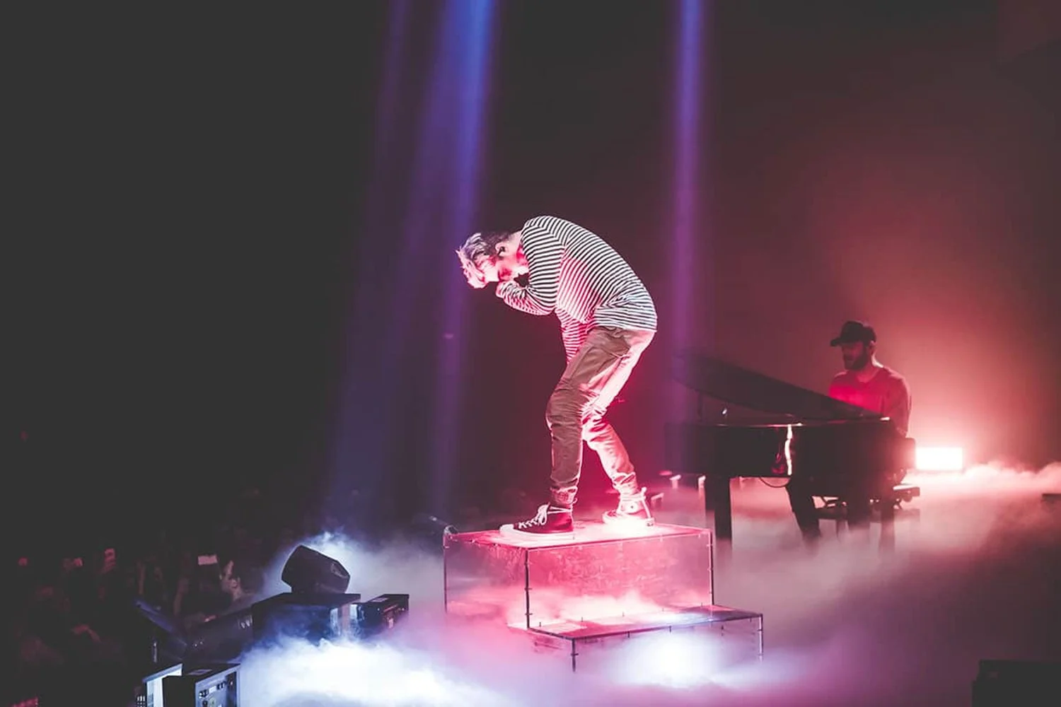 Performer singing on stage with fog and colorful lighting, accompanied by a pianist at a grand piano.