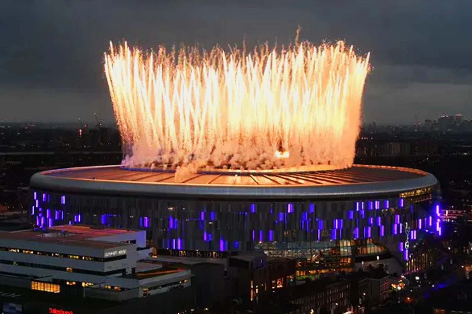 Fireworks display above a circular stadium with purple lighting, during twilight or night.