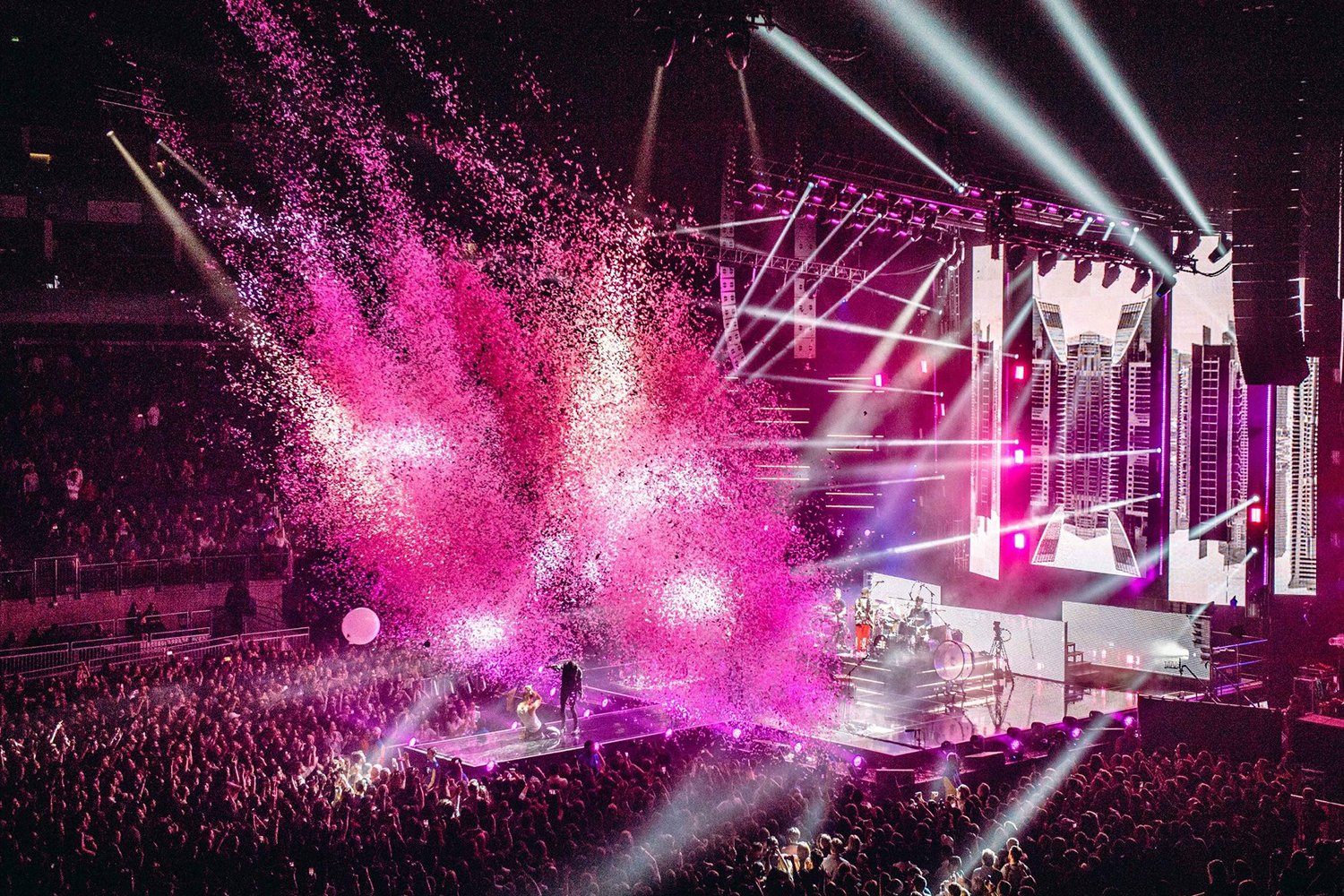 Concert stage with vibrant pink and white lighting, large LED screens, and a crowd of people, with pink confetti falling from above.