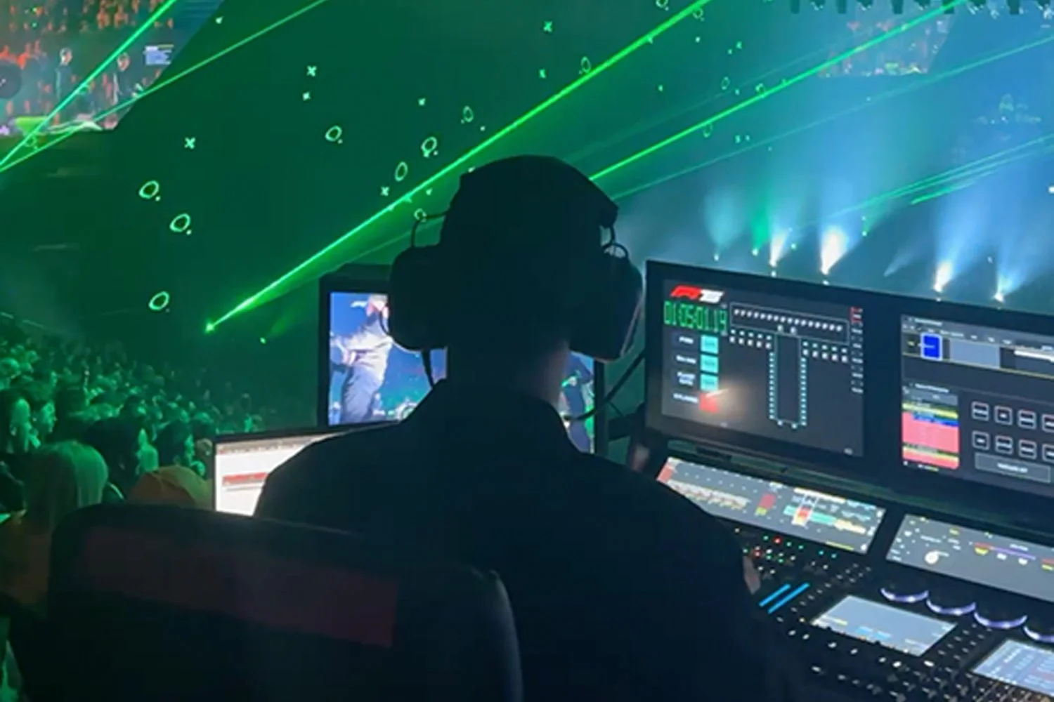 DJ working with electronic music equipment at concert with green laser lights and a large crowd in the background.