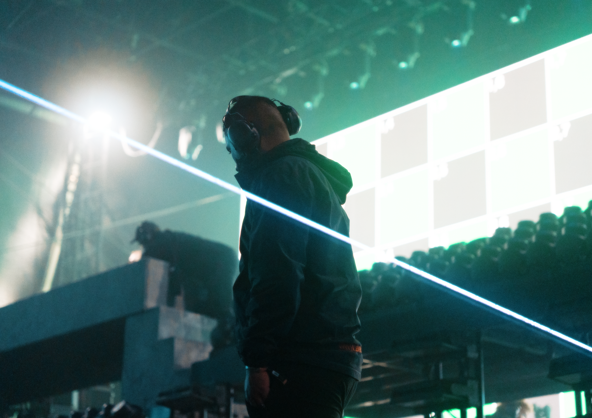 A person wearing headphones standing in front of a large screen with multiple squares and green lines, illuminated by bright lights creating a vibrant atmosphere.