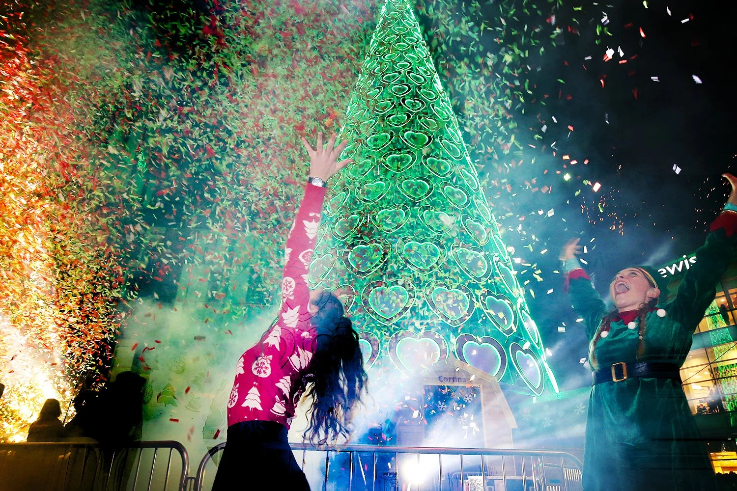Two women celebrating in front of a tall, illuminated Christmas tree with heart-shaped decorations. Festive confetti and lights surround them, and they are joyful with arms raised.