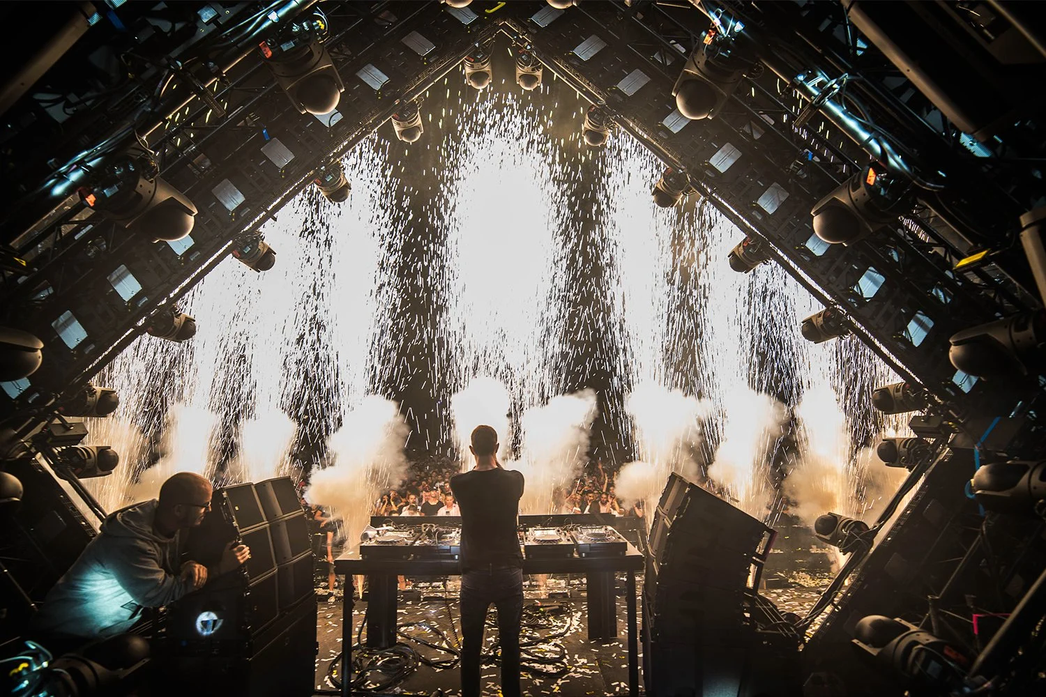 A DJ performs at a concert or music festival with bright fireworks and pyrotechnics in the background, and a crowd of people watching.