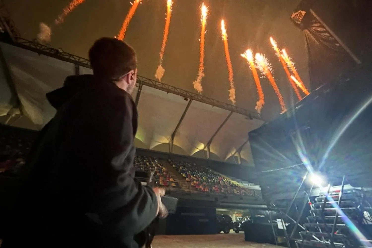 A person wearing a dark hoodie operating sound or DJ equipment inside a large tent with fireworks exploding in the night sky overhead.