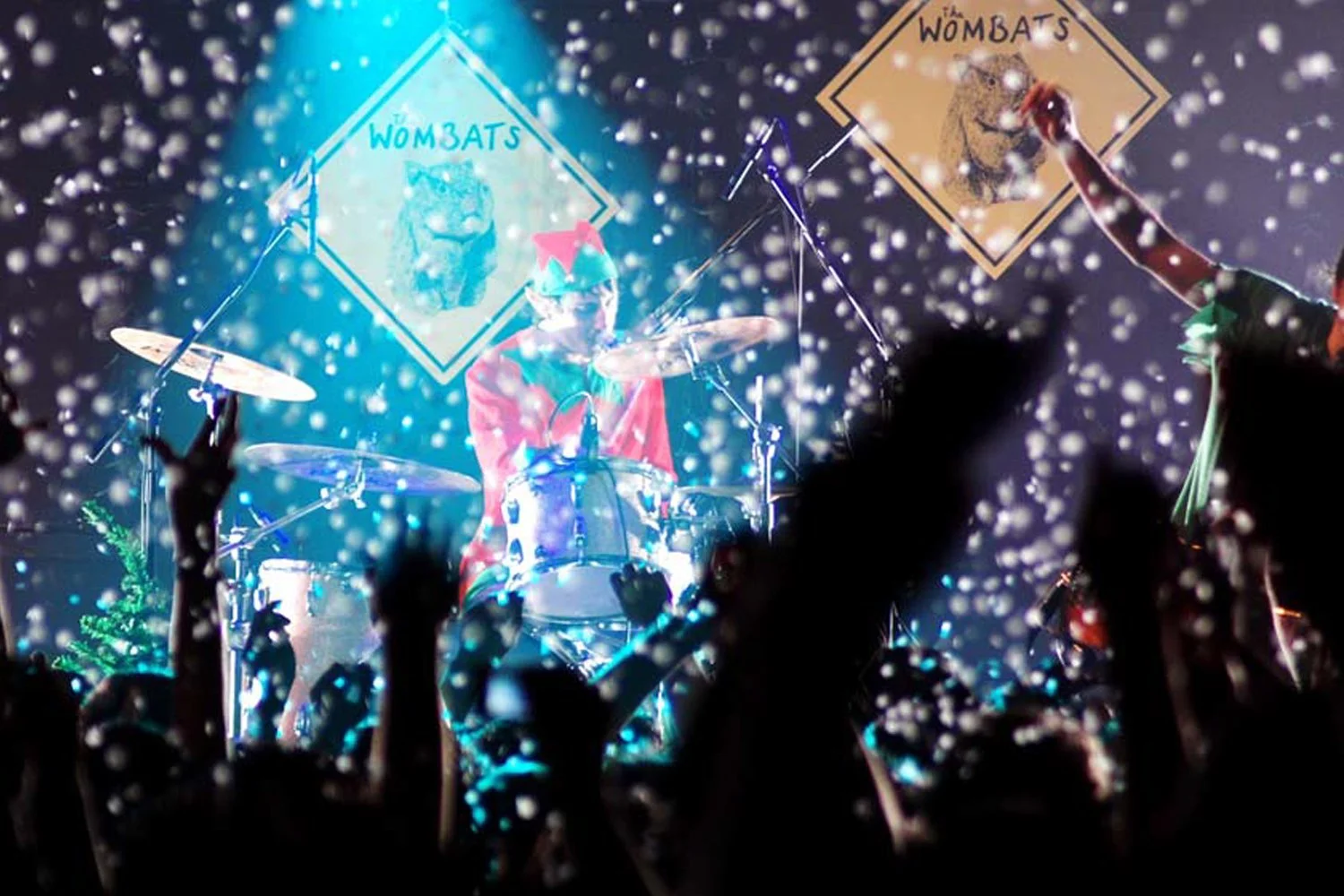 A concert scene with a drummer dressed as an elf, surrounded by falling snow or confetti and illuminated by bright lights. In the background, two diamond-shaped signs display the word 'WOMBATS' with images of wild cats. Audience members have their hands raised.
