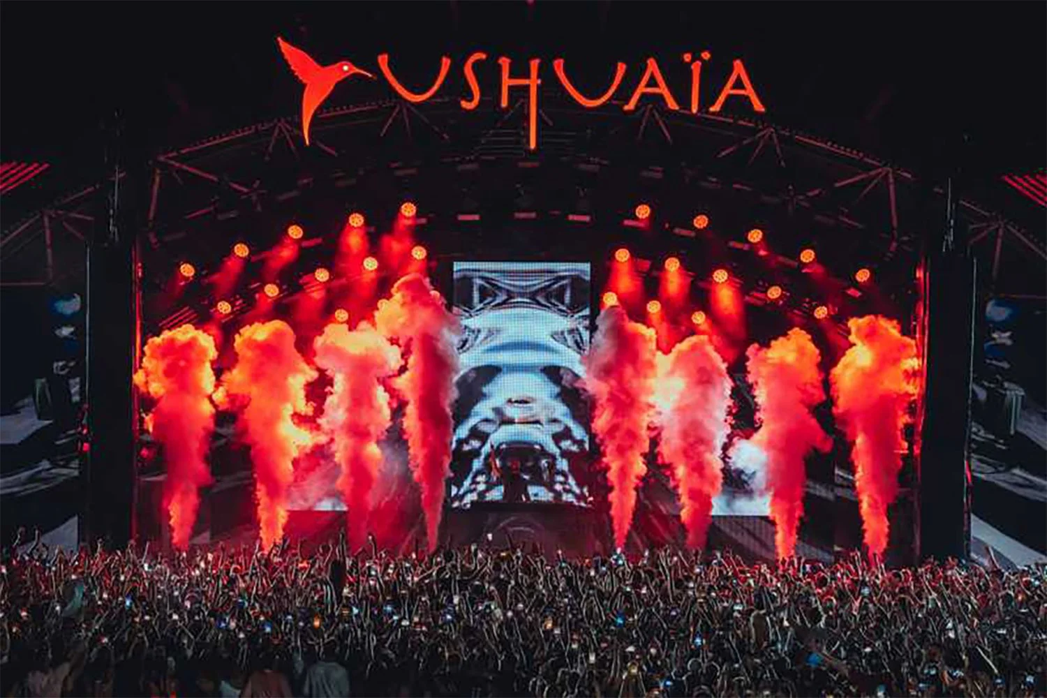 Concert stage with bright red pyrotechnics, large LED screen displaying a panda, and the word "USHUAÏA" in red at the top. Crowds of people are gathered in front of the stage at night.