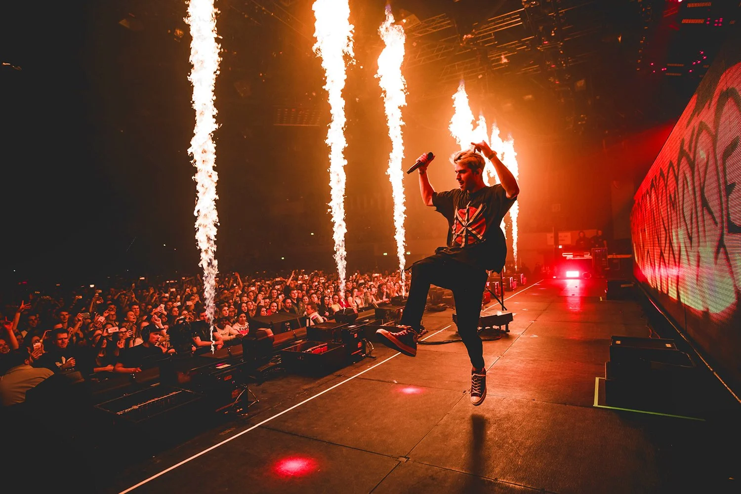 A performer on stage during a concert, jumping with a microphone in hand, with pyrotechnics shooting flames into the air behind them and a large crowd watching.