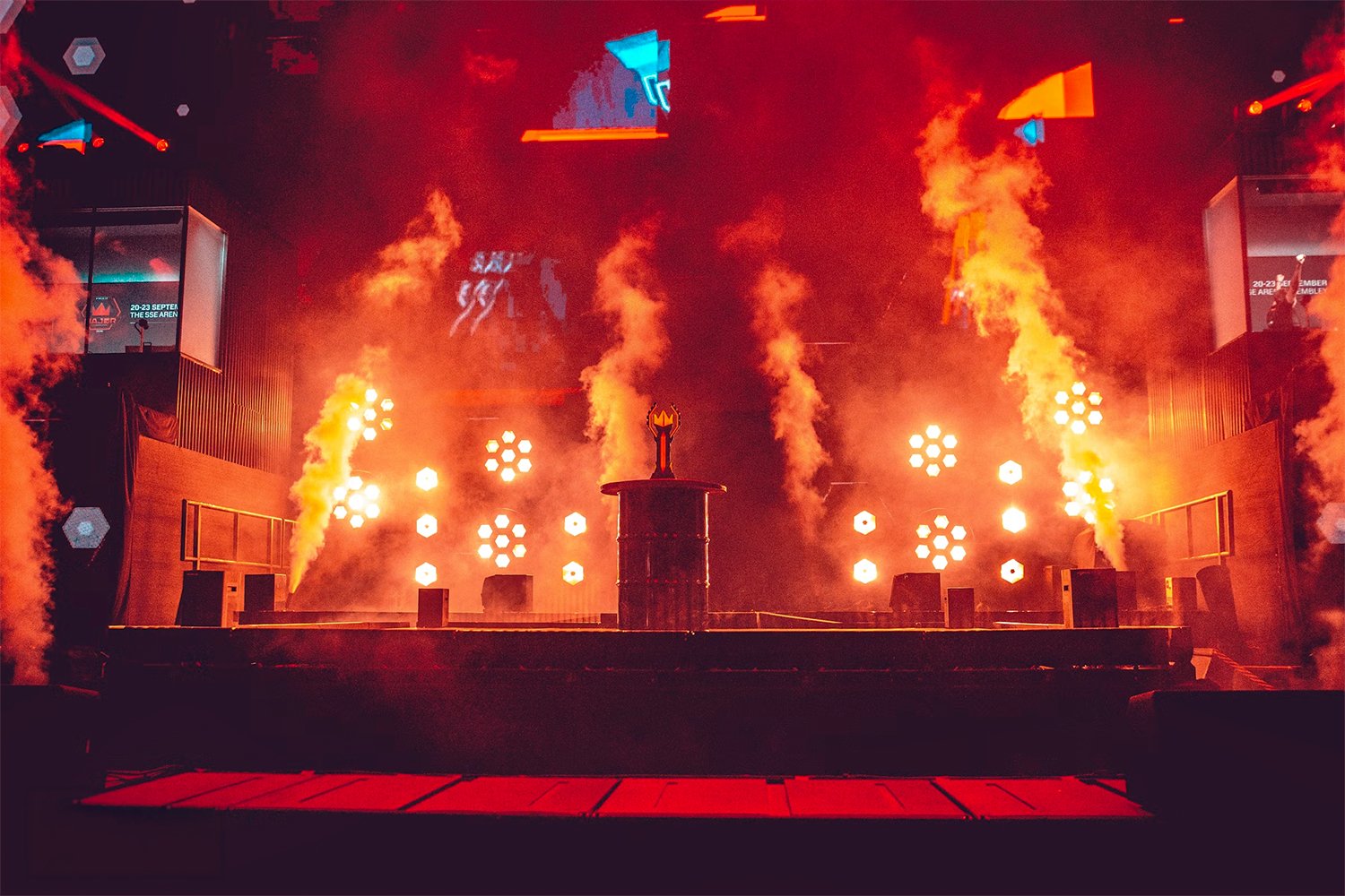 A stage with bright orange and yellow flames and smoke, with a central decorative object on a pedestal. The background features a large screen with colorful geometric shapes and lights, creating an energetic concert or event atmosphere.