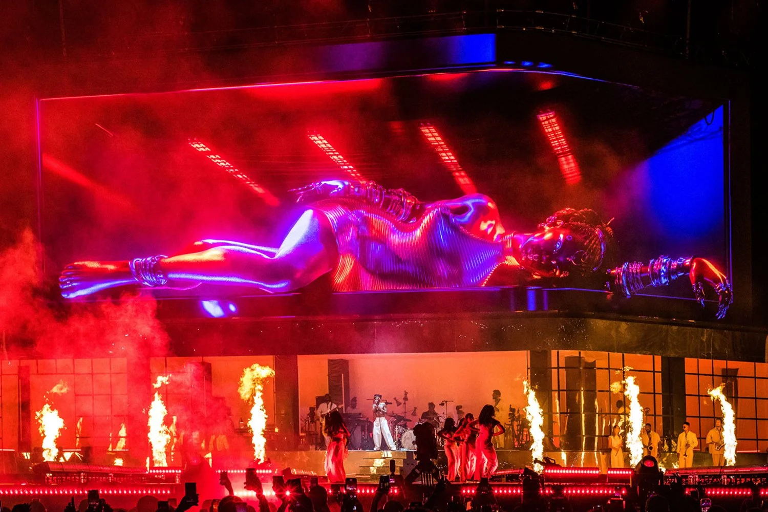 A large digital screen shows a glowing holographic image of a reclining woman with metallic and neon styling, surrounded by red and blue lighting. Below, a stage features performers, dancers, musicians, and pyrotechnics with flames and smoke.